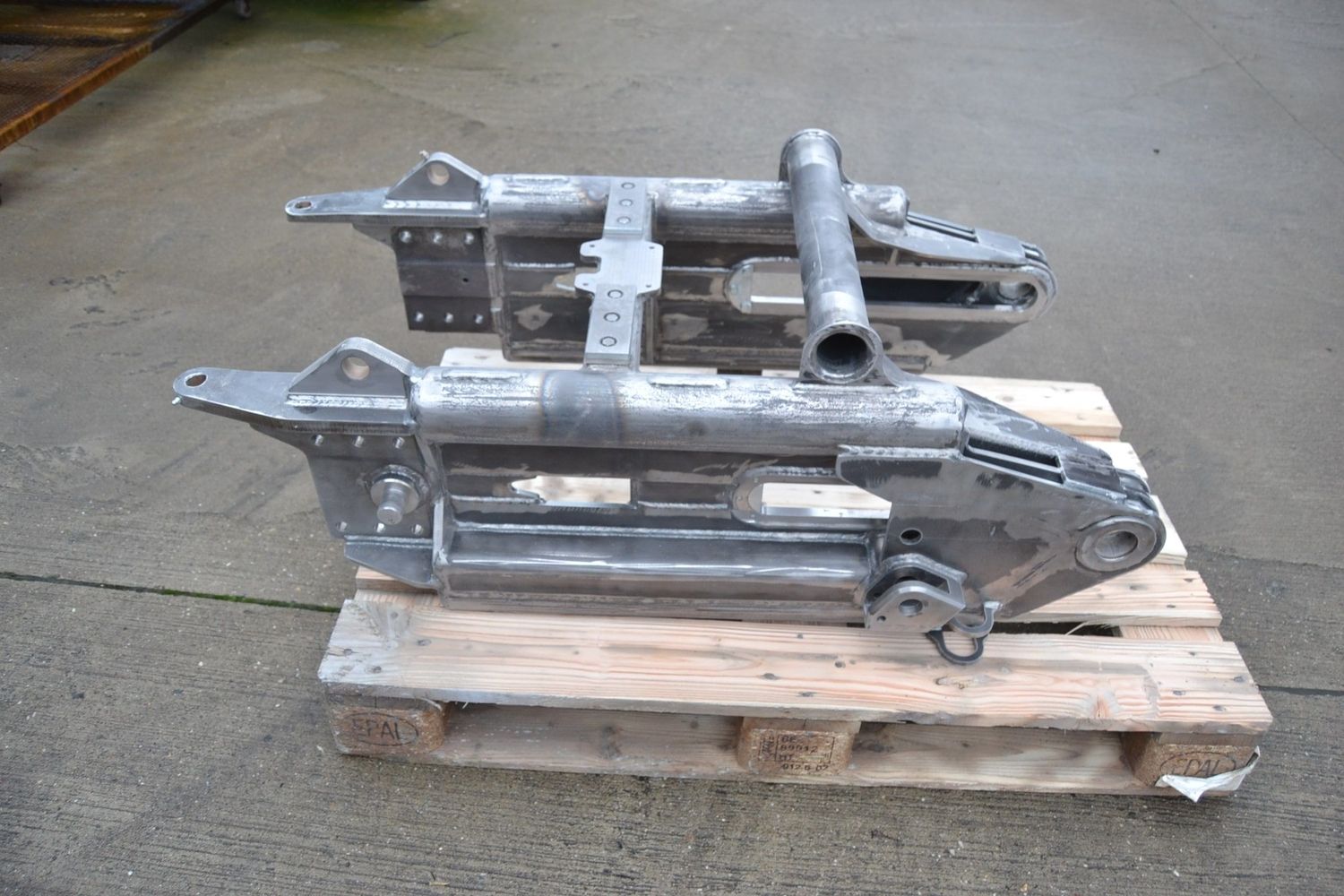 Zwei metallische, industrielle Maschinenteile, die auf einer Holzpalette ruhen und komplizierte Schweißarbeiten und mechanische Konstruktionen zeigen