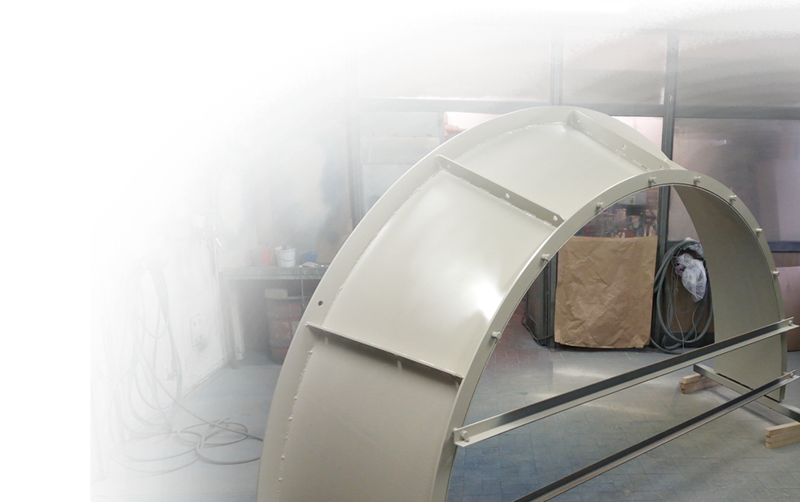 In einer Werkstatt ist eine halbrunde Metallstruktur abgebildet, im Hintergrund sind Farbspritzgeräte und Werkzeuge zu sehen